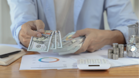 Una persona entrega billetes de 100 dólares mientras realiza cálculos financieros en una mesa con documentos, monedas y una calculadora.