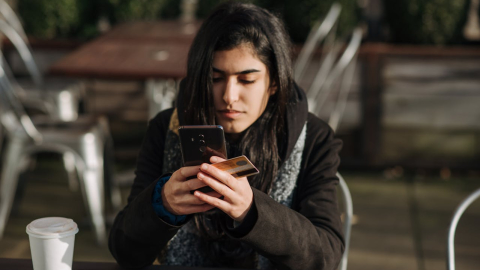 Fotografía referencial de una joven mujer que utiliza una tarjeta de crédito para hacer un pago en su teléfono celular.