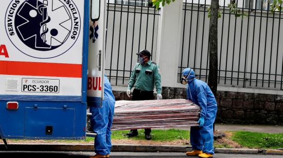 Dos personas de servicios médicos trasladan un ataúd envuelto en plástico.