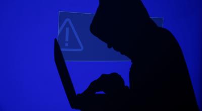 Ilustración. Un hombre encapuchado sostiene una computadora portátil mientras se proyecta sobre él una pantalla azul con un signo de exclamación.